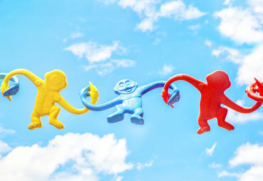 Fotografía de tres changuitos de plasticos colgados entre si, uno es amarillo, otro azul y otro rojo. A fondo se ve un cielo claro con nubes blancas. 