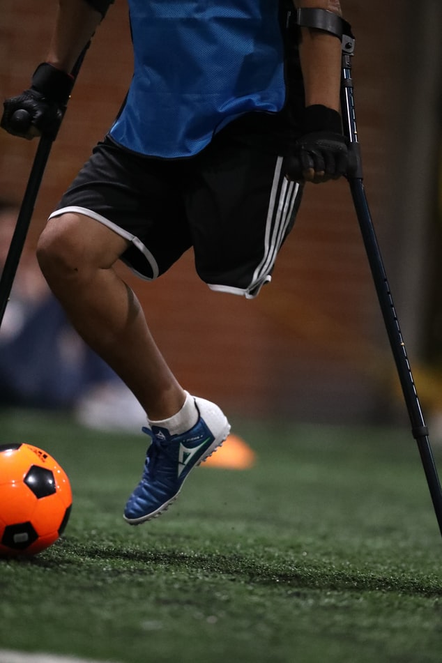 Fotografía del cuerpo de un hombre del pecho hacia el suelo. En una cancha de futbol, usa muletas y le falta una pierna. Patea un balón.