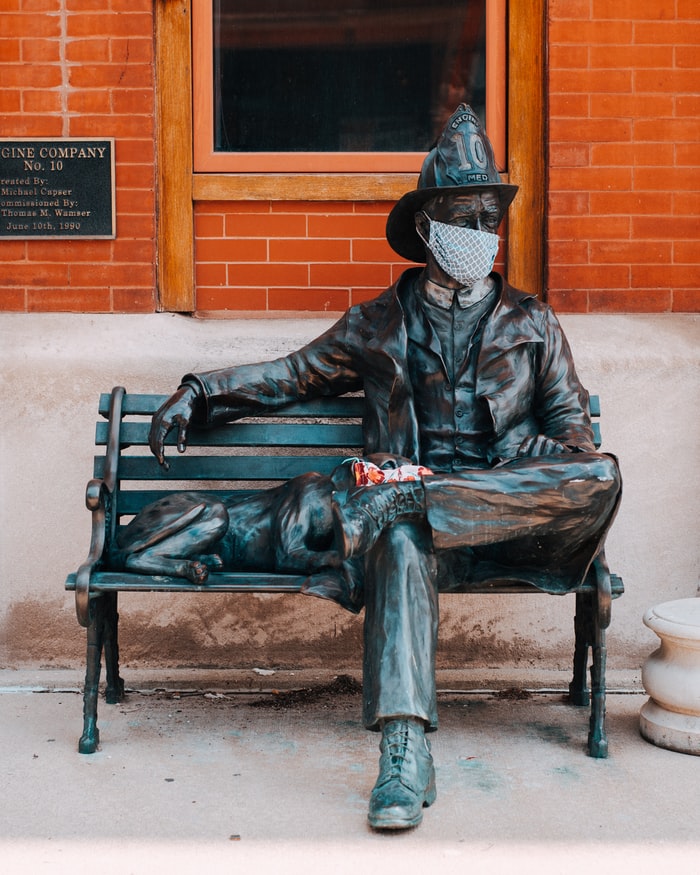 Fotografía de una estatua de bronce de un hombre sentado en una banca que tiene uniforme de policía y a su lado se encuentra un perro. Ambos usan cubrebocas