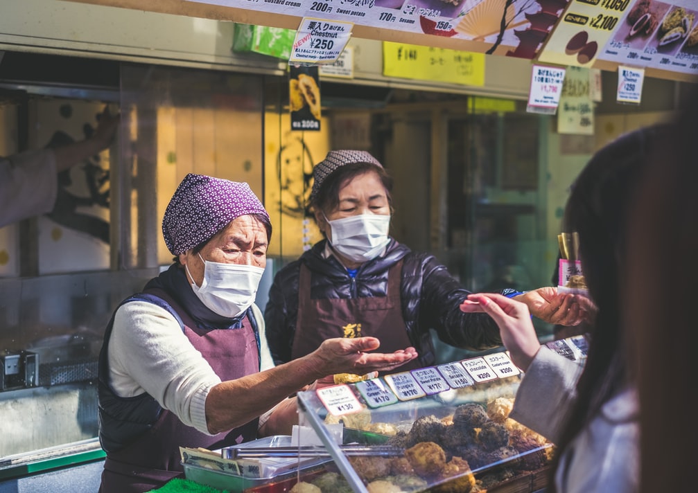 Fotografía de dos mujeres atendiendo en un mercado japones. Ambas mujeres portan un cubrebocas y unas pañoletas en el cabello. 